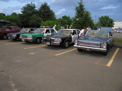 1991 Ford LTD Crown Victoria - row of police cars angled in a striped lot, including a green sheriff car, a black-and-white cruiser, and a blue vintage Ford.