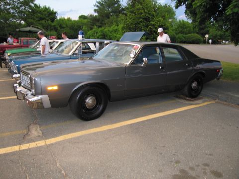 1991 Ford LTD Crown Victoria - gray vintage Dodge police sedan shown in side profile, parked in a striped lot among other police cars with people nearby.