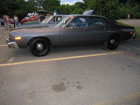 1991 Ford LTD Crown Victoria - gray vintage Dodge police sedan in side profile parked in a yellow-striped lot with people and cars in the background.