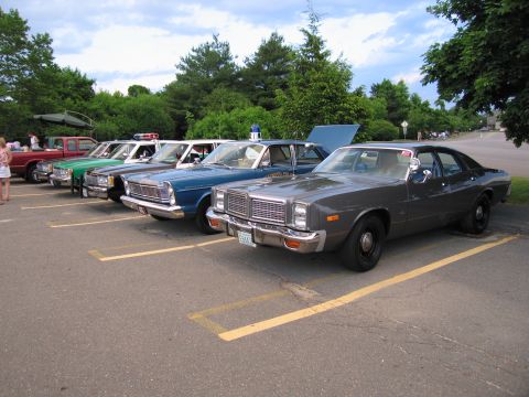 1991 Ford LTD Crown Victoria - row of police cruisers led by a gray Dodge sedan with a green sheriff car and blue vintage Ford behind in a striped lot.
