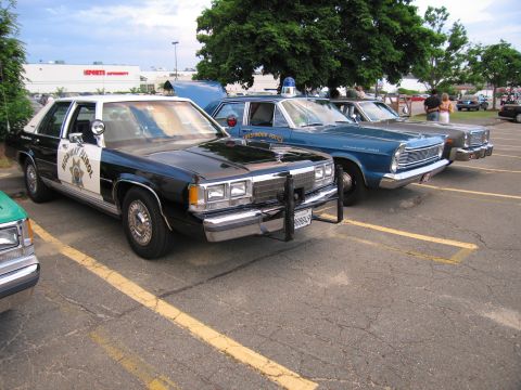 1991 Ford LTD Crown Victoria - black-and-white highway-patrol Ford sedan with star emblem and push bumper parked beside blue vintage police cars in a lot.