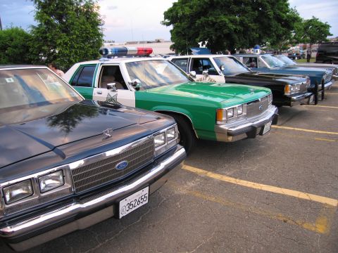 1991 Ford LTD Crown Victoria - row of classic police-livery sedans, including a green-and-white Ford and a black cruiser, parked diagonally in a paved lot.