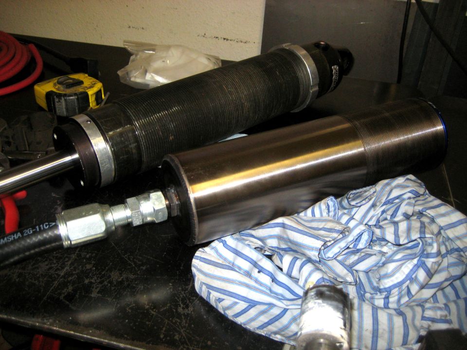Two shock absorbers on a workbench, one with a black ribbed body and the other with a shiny metal body. A hydraulic hose is attached to the metal one