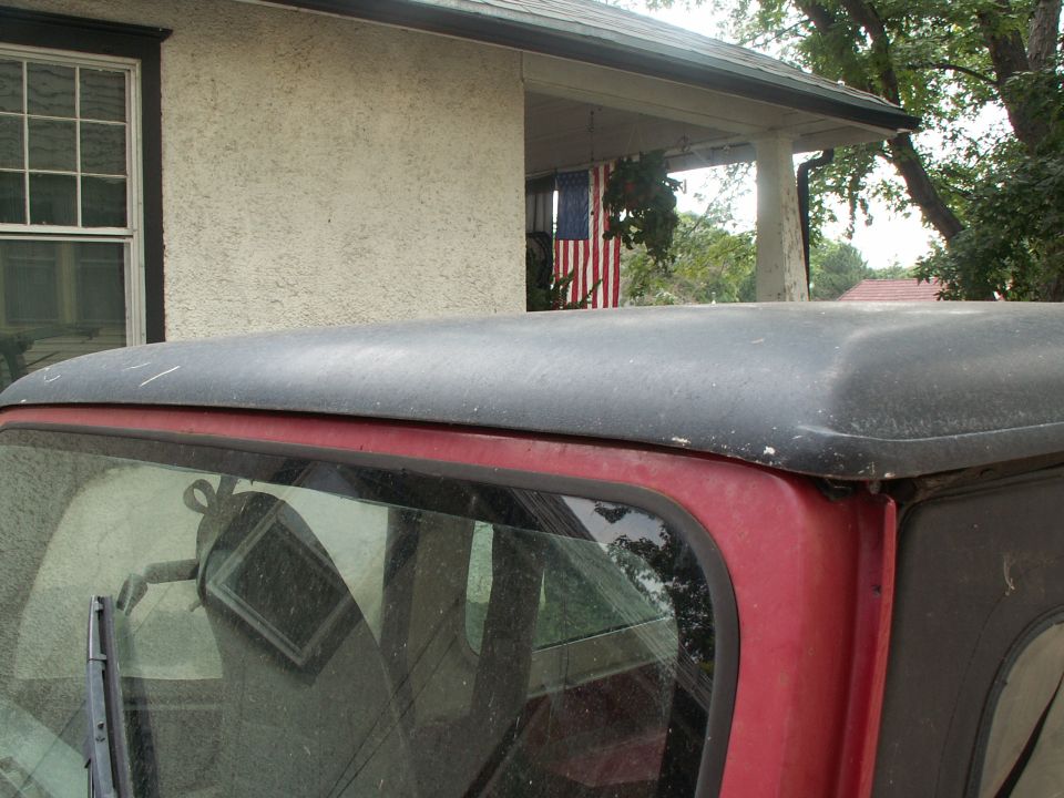 Rear of red Jeep CJ-7 with black hardtop, parked in front of house with American flag hanging on porch