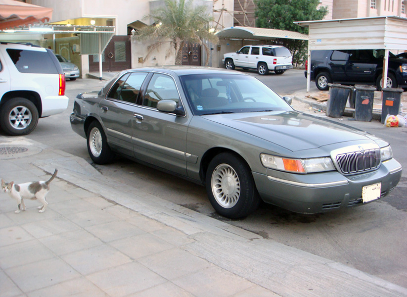 Gray 2001 Mercury Grand Marquis sedan parked on a sidewalk, front quarter view. Visible damage to the front bumper
