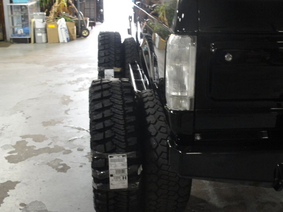 Several large off-road tires stacked next to the rear fender of a black SUV