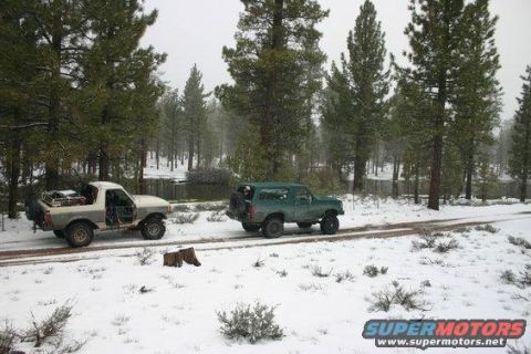 1991 Ford Bronco - Two SUVs drive down a dirt road covered in snow, surrounded by pine trees.