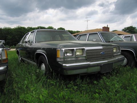 1991 Ford LTD Crown Victoria - A dark green Ford Crown Victoria sits in a grassy field next to other vehicles under a cloudy sky.