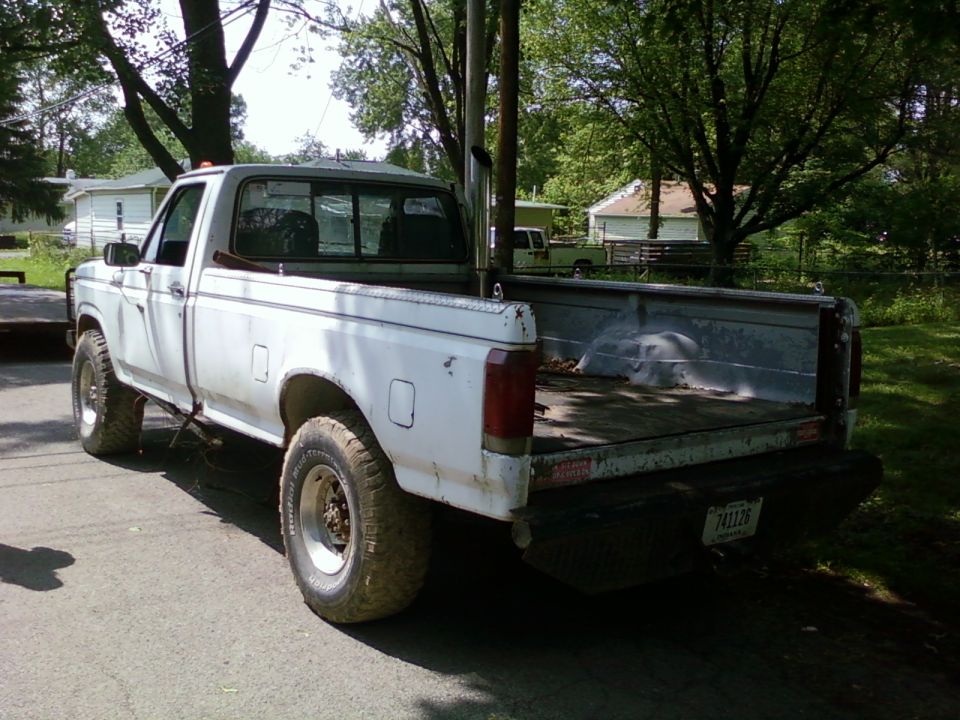 1987 Ford F-150 pickup truck, rear 3/4 view, with large exhaust stack. Another truck is visible in the background