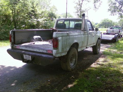1989 Ford Bronco - This weathered pickup truck, likely from the late 1980s, is parked on a dirt road next to a trailer.