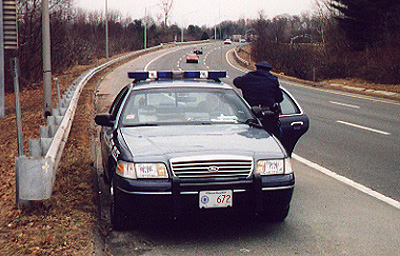 1998 Ford Crown Victoria - A 1998 Ford Crown Victoria police car is parked on the shoulder of a highway, with an officer standing by the open driver's door.