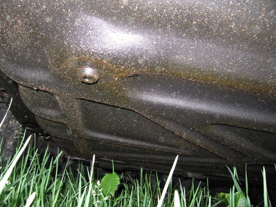 Underside of a car, showing the fuel tank and frame rails, with grass in the foreground