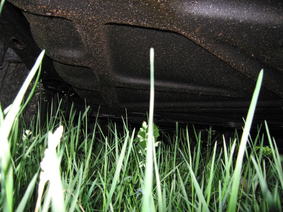 Underside of a dark-colored vehicle chassis, viewed from below through tall green grass