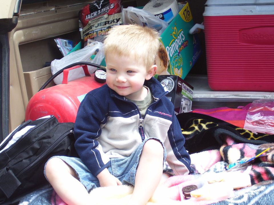 Child sitting in the cargo area of a vehicle, surrounded by items like a red air compressor, diapers, and a cooler