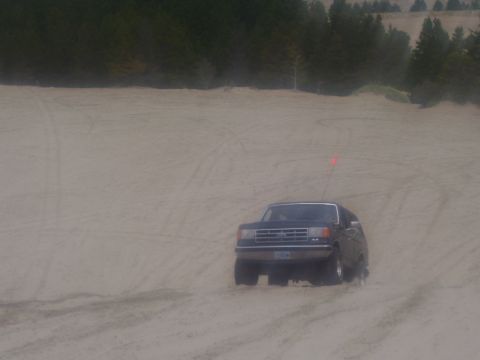 1991 Ford Bronco - A dark SUV navigates sandy terrain, its front grille and a red safety flag clearly visible.