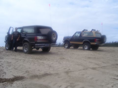 1991 Ford Bronco - Two Ford Broncos are parked on a sandy beach under a cloudy sky. A man stands next to the driver's side door of the vehicle in the foregrou…