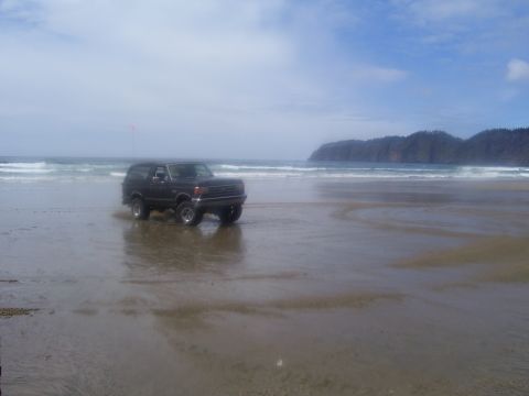 1991 Ford Bronco - A 1991 Ford Bronco navigates the wet sand near the ocean, with waves crashing and a coastal hill in the background.