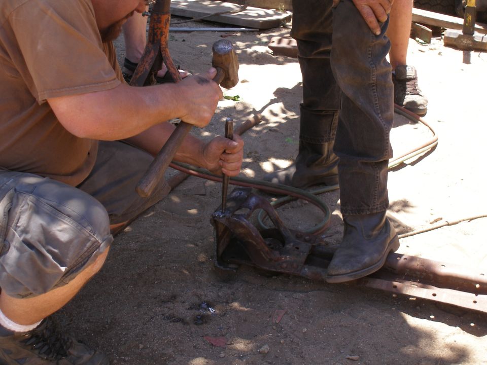 Two people using hammers and chisels on a rusty metal component outdoors