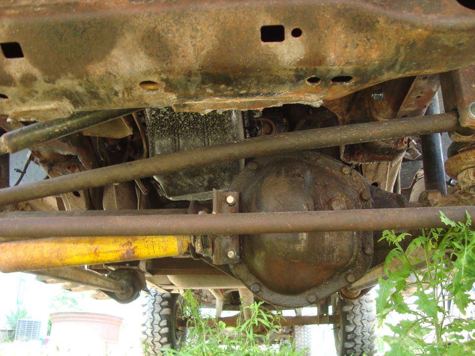 Underside view of a rusty truck frame, showing the differential and suspension components. Green grass is visible below