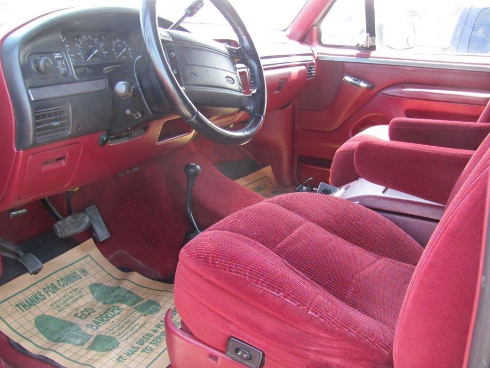 1995 Ford Bronco interior with red velour seats, dashboard, and steering wheel. Floor mat reads 'THANKS FOR COMING IN ECO BARRIER'