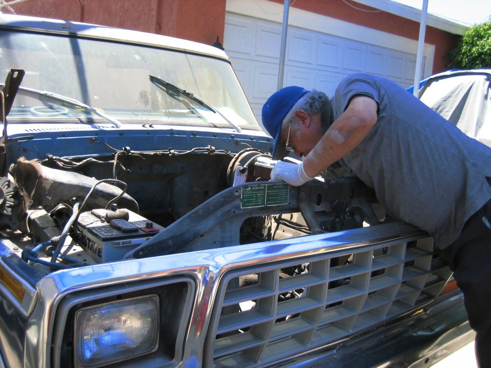 Man in blue cap and gloves works on the engine of a 1979 Ford F-150. The hood is open, showing the battery and engine components