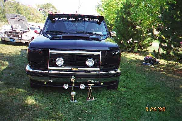 Black Ford truck with 'THE DARK SIDE OF THE MOON' banner on windshield, trophies in front. Another car with hood up in background
