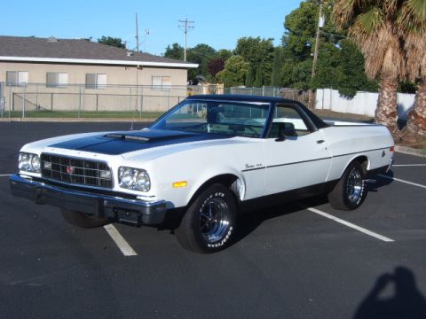 1973 Ford Ranchero - A white 1973 Ford Ranchero with a black hood and roof is parked in an asphalt lot with a building and trees in the background.