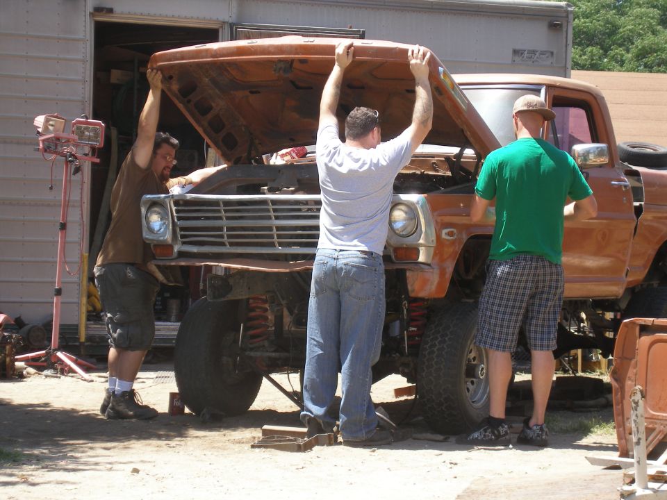 Three guys working on the front of an orange pickup truck with the hood up
