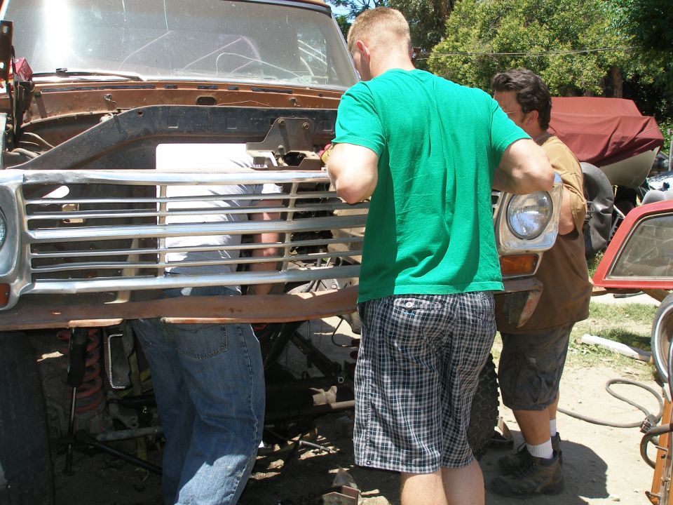 Rusty truck grille with horizontal chrome bars, being worked on by two men