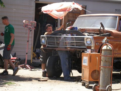 1969 Ford F-250 - Two men work on a truck's engine bay, using an umbrella for shade. Welding equipment is set up next to the vehicle.
