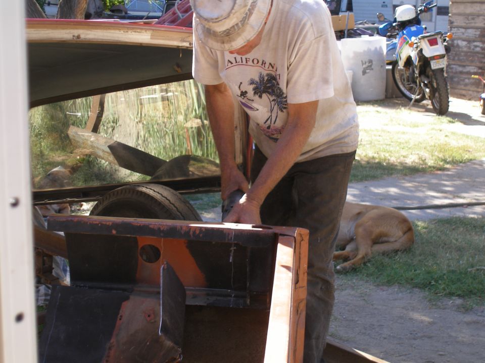 Man grinding on a rusty vehicle frame outdoors. Dog sleeping nearby. Motorcycle in background