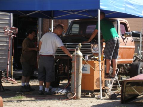 1969 Ford F-250 - Men work on the front end of an orange pickup truck under a blue canopy, with welding equipment nearby.