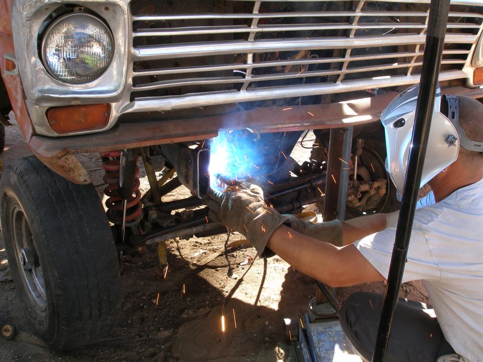 Ford truck front end with chrome grille and round headlight, being welded by a person in protective gear