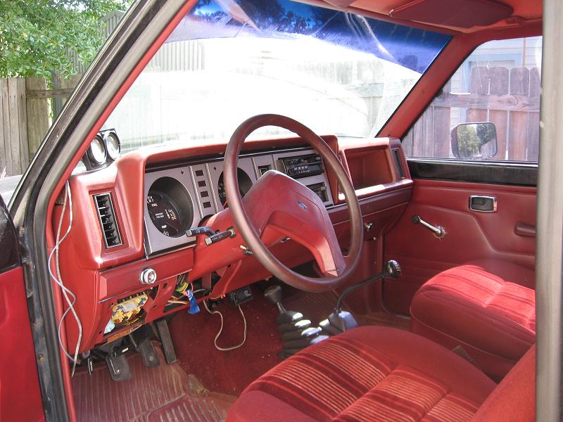 Interior of a red truck, showing the dashboard, steering wheel, and seats. Wires are visible under the dash