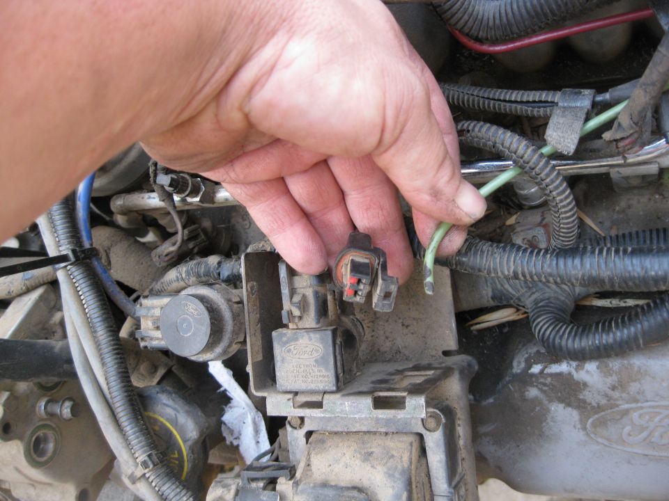 Hand disconnecting a green wire from an engine wiring harness connector. Ford logo visible on part