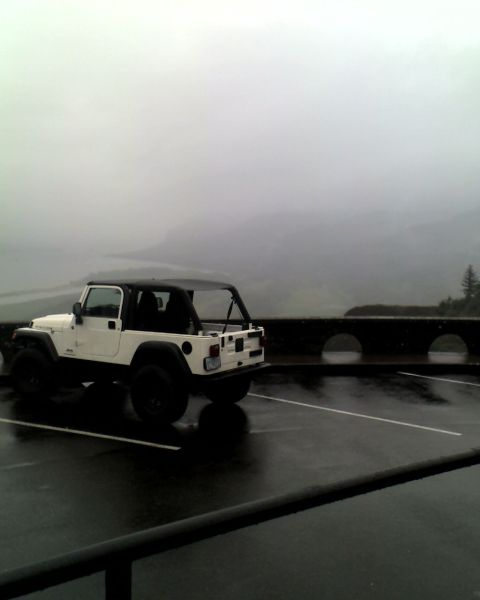 2005 Jeep Wrangler - A white Jeep Wrangler is parked in a wet parking lot with a scenic, misty mountain landscape in the background.