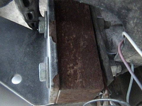 1995 Ford Bronco - Close-up underside shot of a rusty rectangular steel spacer block sandwiched between a bumper bracket and the frame rail, held by large hex…