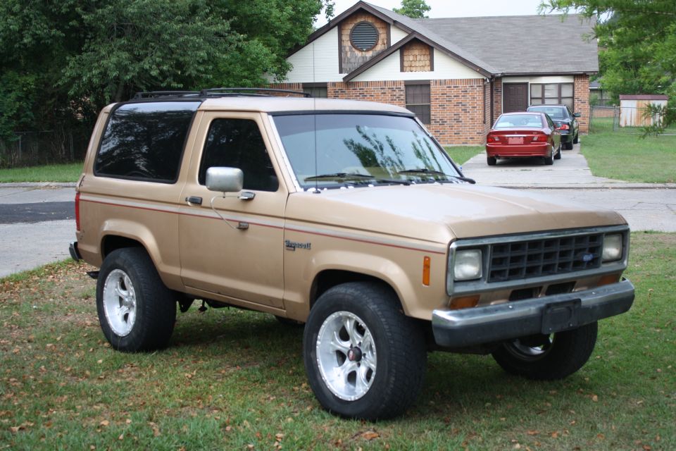 Tan Ford Bronco II with aftermarket wheels parked on grass in front of a house