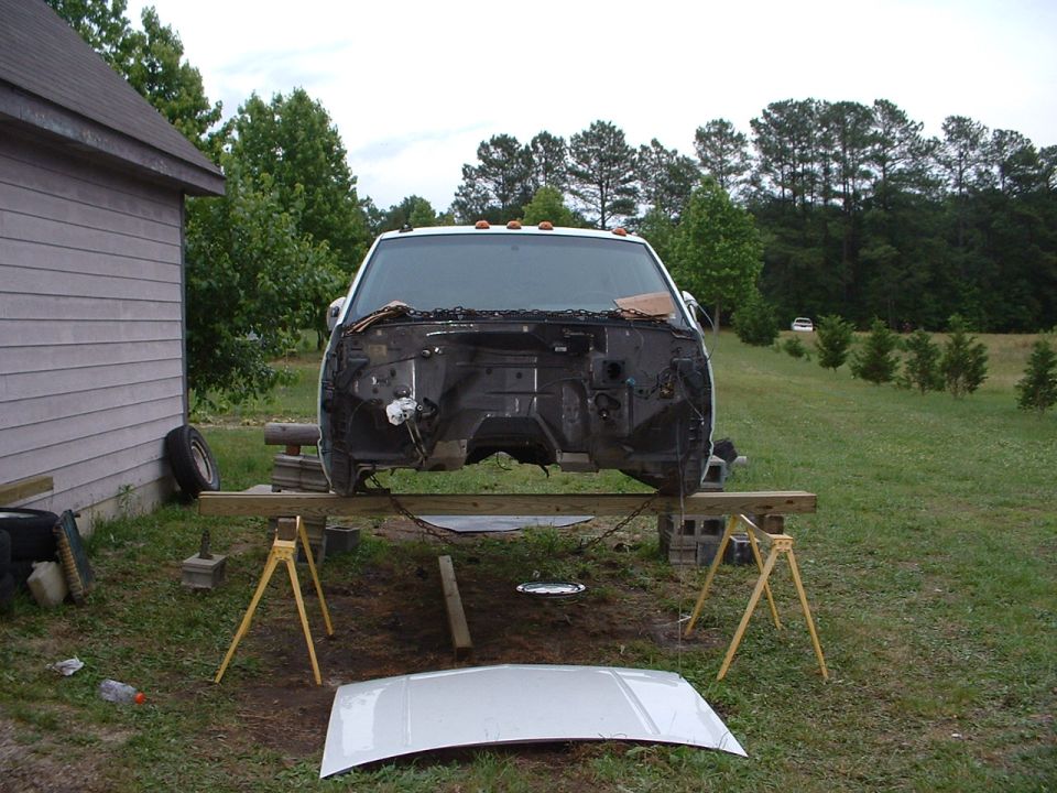 White truck body shell on a beam and sawhorses with stripped front end and hood on the grass
