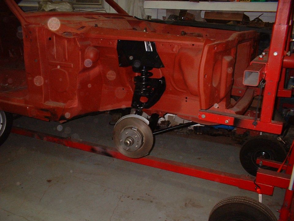 Stripped classic car front end in red primer on a rotisserie showing suspension and brake rotor