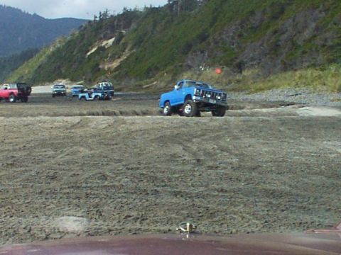 1980 Ford Bronco - Several trucks are parked on a sandy beach with a hill in the background.
