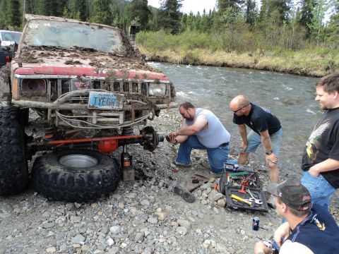1990 Chevrolet Suburban - Several men work on a heavily mud-caked off-road vehicle next to a flowing river.