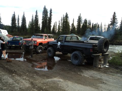 1990 Chevrolet Suburban - Several lifted trucks are parked in a muddy, wooded area near a river.