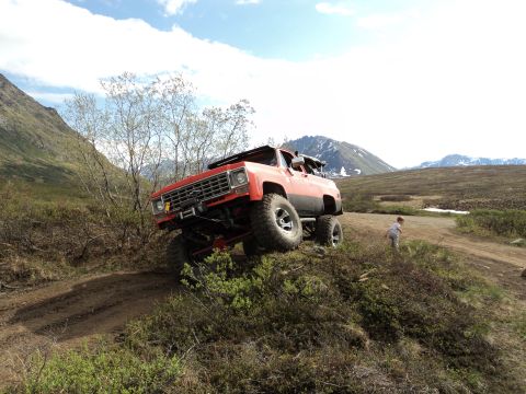 1990 Chevrolet Suburban - A modified red truck navigates a challenging off-road terrain with a mountainous landscape.