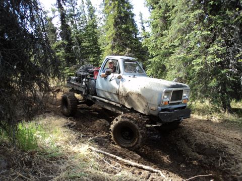 1990 Chevrolet Suburban - A lifted pickup truck with oversized tires drives through a muddy forest path.