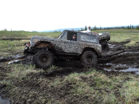 1990 Chevrolet Suburban - Two off-road vehicles are stuck in a muddy field, with people visible inside the front vehicle.