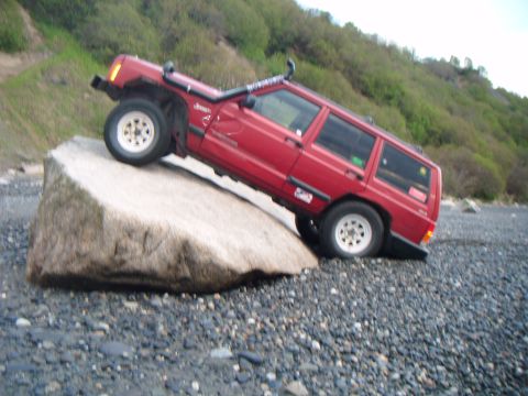 1998 Jeep Cherokee - A red 1998 Jeep Cherokee is shown navigating a large rock on a gravelly beach.