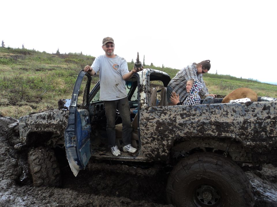 Mud-caked truck with open driver's door, people in cab and bed, dog in bed