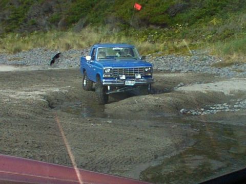 1980 Ford Bronco - A blue pickup truck navigates a challenging off-road terrain with a red flag visible in the distance.
