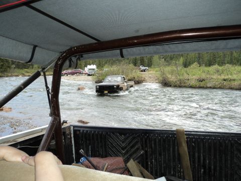 1990 Chevrolet Suburban - Several vehicles are seen crossing a river in a wooded area, with campers parked on the shore.
